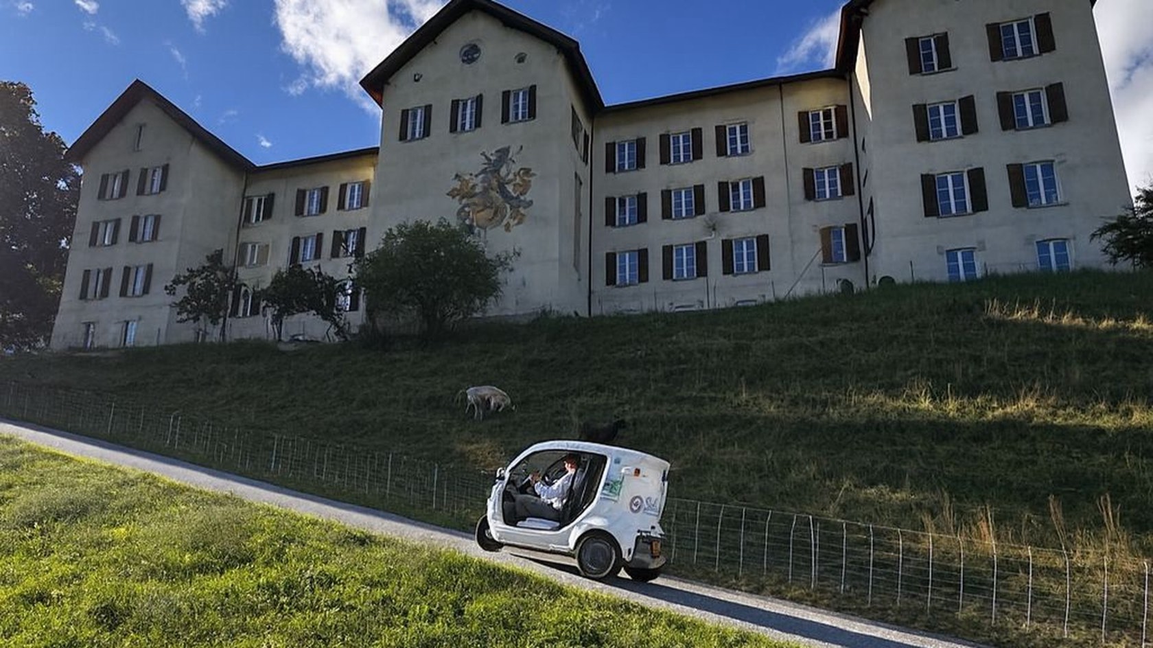 Weisses eMiMo-Elektrofahrzeug auf der Strasse unterhalb des Klosters Löwenberg in der Surselva, Schafe an der Wiese.